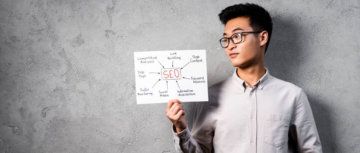 A man with glasses is holding a piece of paper that has "SEO" written in the center, surrounded by related terms like "Link Building," "Page Content," "Keyword Research," and more. He is standing against a gray wall.