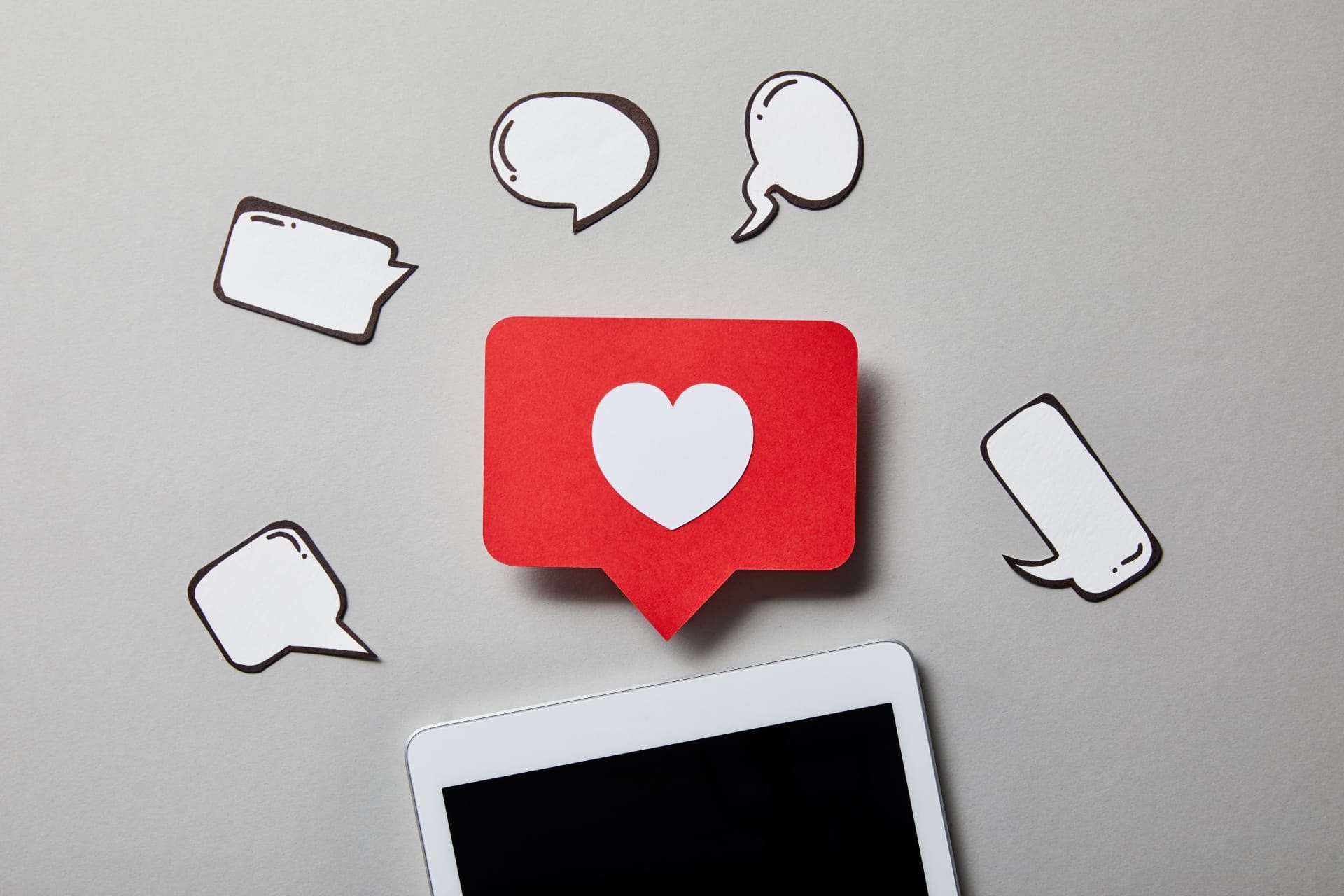 A red speech bubble with a white heart icon and several blank speech bubbles surround a tablet with a blank screen on a gray background, perfectly illustrating the connectivity and engagement central to effective social media marketing.